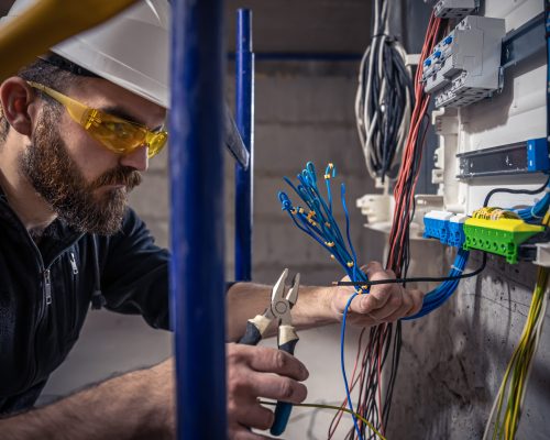 A male electrician works in a switchboard with an electrical connecting cable, connects the equipment with tools.